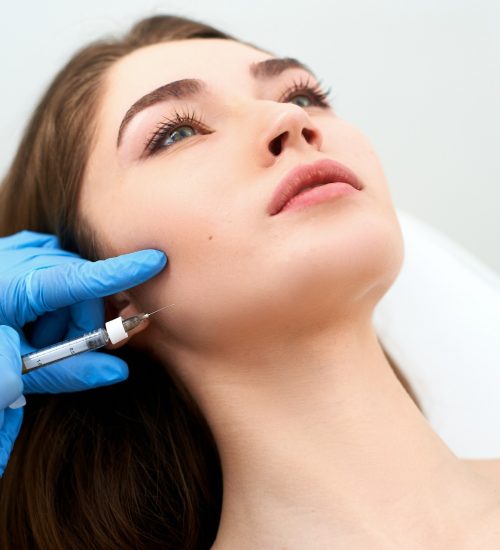 Beautician doctor with filler syringe making injection to jowls. Masseter lines reduction and face contouring therapy. Anti-aging treatment and face lift in cosmetology clinic. Patient lying on medical chair.